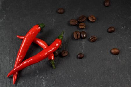 Abstractional design of 3 red hot chili in the foreground and scattered maricevich coffee beans in the background. On the black background of natural stone. Side view. Close-upの写真素材