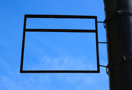 An empty old and dirty rectangular metal frame or framework hanging on a metallic black pillar against a blue sky background with copy space. A blank rectangular sign framing a blue sky background.の写真素材