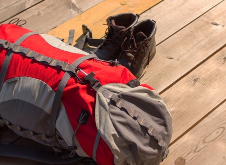 Red tourist backpack and dusty old hiking boots on a background of rough wooden boards on a sunny day. Ideal concept background for wanderlust of a tourists and travelers with space for text.の写真素材