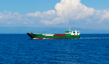 Loaded cargo ferry in the open sea with blue water on the background of the coastline and cloudsの写真素材