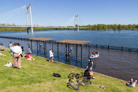 Krasnoyarsk, Russia - June 1, 2021: people are resting on the lawn and swing on a swing against the background of the flooded city embankment on a sunny day. Natural disasterのeditorial素材