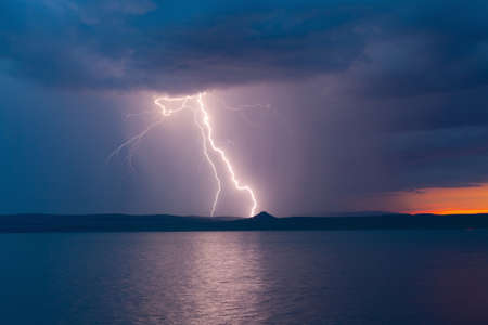 Bright lightning strike during a thunderstorm in the twilight stormy sky over the water of the lake