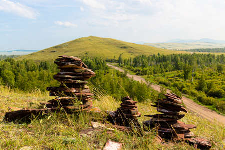 Three cairns from reddish balanced stones of Devonian sandstone on the top of hill against summer mountain landscape. Sunduki mountain range in Khakassia, Russiaの写真素材