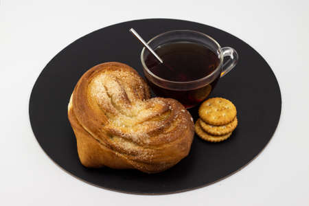 Still life with yeast sweet heart shaped bun with a cup of black tea and crispy crackers on a black tray and white background. Russian tea drinking concept. Top viewの写真素材