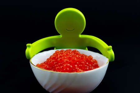Smiling bright green plastic toy man hugs a white ceramic bowl fully filled with red caviar on black...