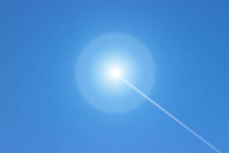 White fluffy condensing trail from the plane ends in the sun against a bright blue sky. Solar flare...