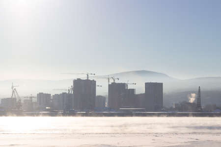 Silhouettes of buildings and cranes through winter fog on construction site. Morning landscape of thick fog over the river in cold winter day. Cityscape with Yenisey river in Krasnoyarsk, Russiaの写真素材