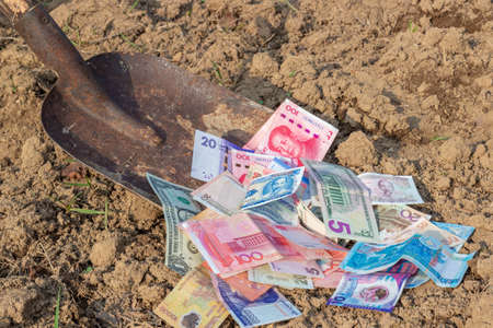 Rowing money with an old rusty coal shovel. Shoveling world currencies banknotes from ground. Money of the different countries on soil.の写真素材