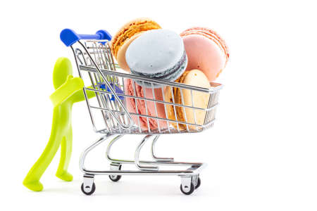 Green plastic toy abstract figure of man pushing full shopping cart with macaroon cookies isolated on white background. Greeting card for Valentine's day. Ideas for shopping in sweet-shop and cafeの写真素材