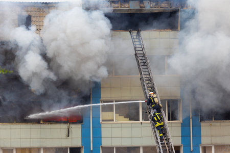 Firemen on telescopic ladder extinguishing strong fire with from a fire fighting firehose nozzle in rescue operation.Thick gray smoke and fire from the windows without glass of industrial warehouse.の写真素材