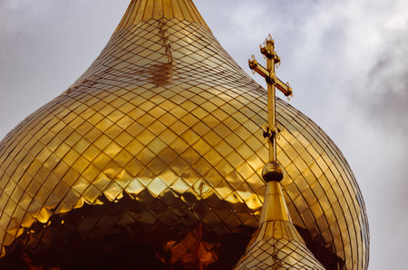 Golden cross on the dome of the church against the background of the big golden dome of the orthodox church against gray cloudy sky. christian religion backgroundの写真素材