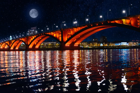 Night landscape with full moon over the illuminated of Communal Bridge in Krasnoyarsk, Russia. View of the Yenisey River at starlight night. Reflection of lighting lights in the rippled water surfaceの写真素材