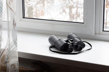 Black binoculars on the white windowsill near a window. The concept of watching and peeping from the window of the houseの写真素材
