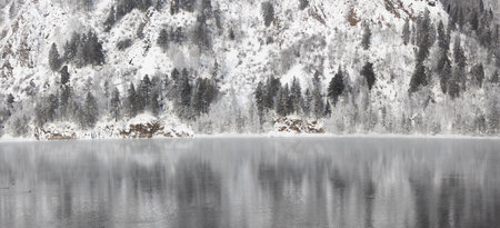 Panoramic landscape of slow river and winter forest covered with frost on riverbank. Pine trees on a steep mountain slope. Siberian winter landscape with river and forestの写真素材