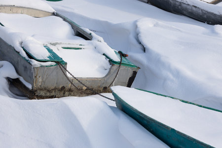 Boats chained together in a snowdrift in the winter season. Metal boats covered with snow on the shoreの写真素材