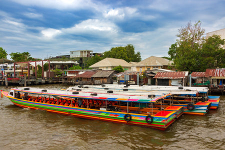 Bangkok, Thailand - December 29, 2023: Empty colorful wooden tourist boats against the background of collapsing wooden residential slums on stilts on the Chao Phraya River in Bangkok, Thailandのeditorial素材