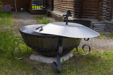 A giant metal vat for holy water stands outside on the grounds of an Orthodox Christianity church. The lid of the vat is decorated with a religious cross.の写真素材