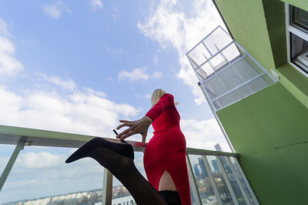 Young beautiful woman in a red dress and high heels on the balcony of an apartmentの写真素材