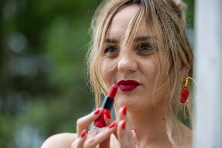 A photo of a woman applying red lipstick with matching red nails, standing outdoors. She is wearing gold earrings with red accents, and the background is filled with greeneryの写真素材
