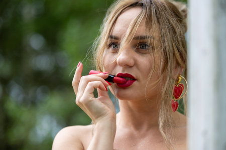 A photo of a woman applying red lipstick with matching red nails, standing outdoors. She is wearing...