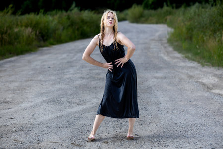 Beautiful blonde barefoot girl in a black dress on a gravel roadの写真素材