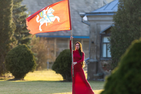 An elegant brunette in a red dress proudly poses, holding the wind-swept historical flag of Lithuaniaの写真素材