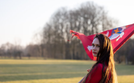 An elegant brunette in a red dress proudly poses, holding the wind-swept historical flag of...