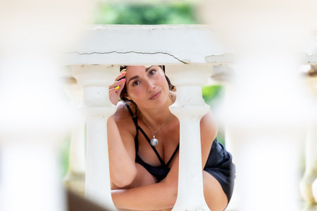A confident young woman poses gracefully between white classical balustrades, playfully peeking through the architectural columns. Her expression is soft and engaging, with a slight smile that conveys charm and openness. The composition blends elegance, geometry, and personality â ideal for themes of femininity, architectural detail, freedom, natural beauty, and spontaneous portraitureの写真素材