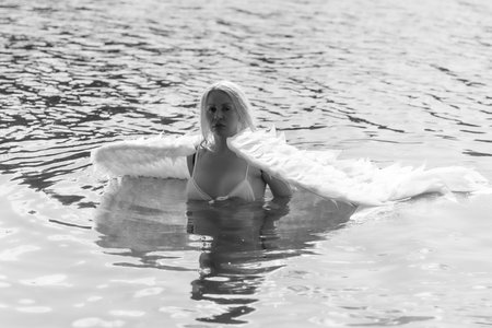 A graceful woman with angel-like wings standing in a calm lake, surrounded by golden light and reeds. This poetic image blends nature, femininity, and quiet emotion, evoking serenity, transformation, and mythological purity.の写真素材