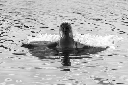 A graceful woman with angel-like wings standing in a calm lake, surrounded by golden light and reeds. This poetic image blends nature, femininity, and quiet emotion, evoking serenity, transformation, and mythological purity.の写真素材