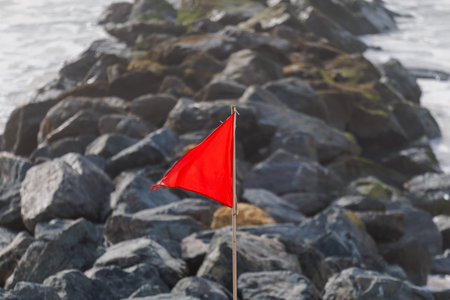 A red warning flag waves in the wind by a rocky breakwater in Lacanau, France. It signals dangerous...