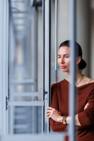 Calm thoughtful woman standing by the large window, wearing a brown sweater and jeans, looking...