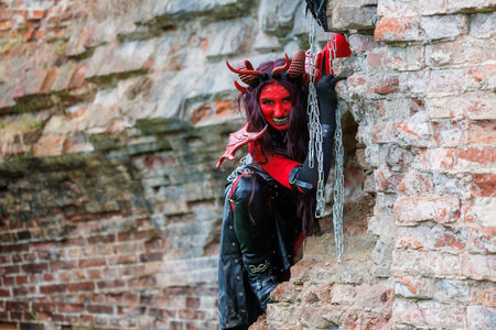 A woman in demonic cosplay with red body paint, horns and chains poses against an old brick wall....