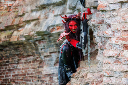 A woman in demonic cosplay with red body paint, horns and chains poses against an old brick wall....