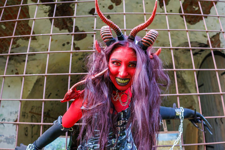 A woman in demonic cosplay with red body paint, horns and chains poses behind metal bars in an...