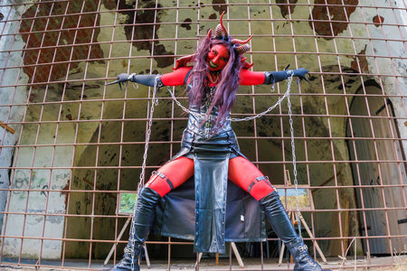 A woman in demonic cosplay with red body paint, horns and chains poses behind metal bars in an abandoned dungeon-like setting. Dark fantasy character in gothic costume and dramatic makeup expressing power and intensityの写真素材