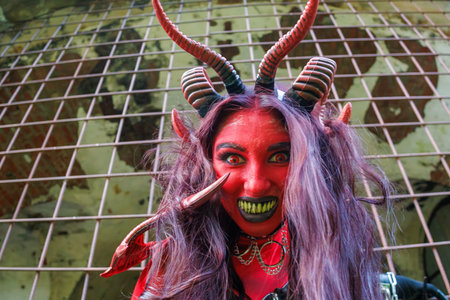 A woman in demonic cosplay with red body paint, horns and chains poses behind metal bars in an abandoned dungeon-like setting. Dark fantasy character in gothic costume and dramatic makeup expressing power and intensityの写真素材