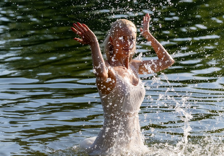 Joyful young woman in a white summer dress enjoying playful water splashes in a natural lake setting on a sunny day. Energetic motion captured in mid-air with sparkling water drops, creating a sense of freedom, freshness, and happiness in nature. Perfect for concepts of summer, vitality, relaxation, and outdoor lifestyle.の写真素材