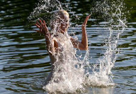 Joyful young woman in a white summer dress enjoying playful water splashes in a natural lake setting on a sunny day. Energetic motion captured in mid-air with sparkling water drops, creating a sense of freedom, freshness, and happiness in nature. Perfect for concepts of summer, vitality, relaxation, and outdoor lifestyle.の写真素材
