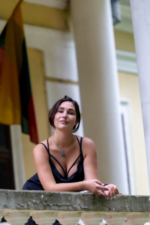 A graceful woman in a sleek black dress stands on the balcony of a historic manor, framed by classical columns. Her elegance and confident pose contrast beautifully with the weathered architecture behind her. A visual story of style, heritage, and national identity.の写真素材
