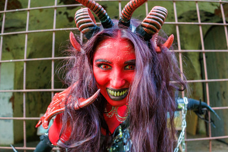 A woman in demonic cosplay with red body paint, horns and chains poses behind metal bars in an...