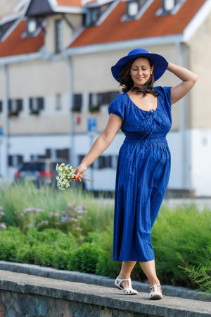 A woman in a blue summer dress walks outdoors on a warm day, holding flowers and a wide-brimmed hat. A cheerful, carefree lifestyle moment in an urban environment, full of light and natural emotionの写真素材