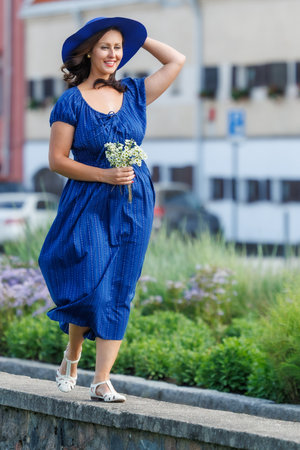 A woman in a blue summer dress walks outdoors on a warm day, holding flowers and a wide-brimmed hat. A cheerful, carefree lifestyle moment in an urban environment, full of light and natural emotionの写真素材