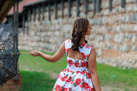 A smiling woman in a floral summer dress stands outdoors with a lace parasol, enjoying a warm and sunny day. A cheerful lifestyle moment with elegance and classic feminine charmの写真素材