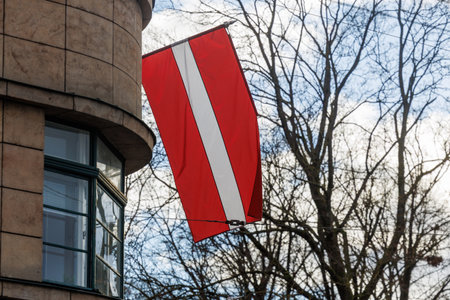 Latvian national flags displayed on building facades in an urban environment, waving in natural light. Red-white-red colors shown in various city streets and architectural settingsの写真素材