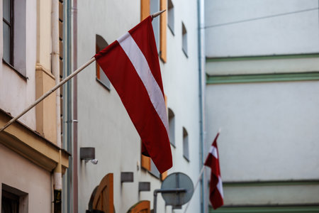 Latvian national flags displayed on building facades in an urban environment, waving in natural light. Red-white-red colors shown in various city streets and architectural settingsの写真素材