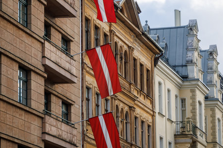Latvian national flags displayed on building facades in an urban environment, waving in natural light. Red-white-red colors shown in various city streets and architectural settingsの写真素材