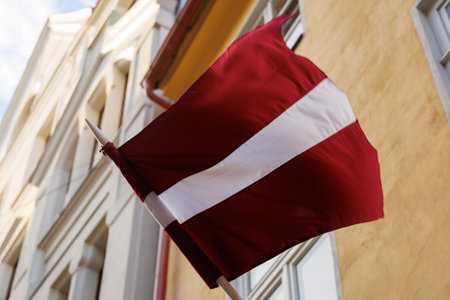 Latvian national flags displayed on building facades in an urban environment, waving in natural light. Red-white-red colors shown in various city streets and architectural settingsの写真素材
