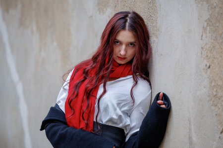 Portrait of a young woman with long hair and a red scarf standing by a textured wall. Natural outdoor light and neutral tones create a calm, stylish and expressive mood suitable for lifestyle themesの写真素材