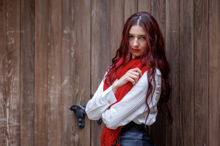 Young woman posing in front of a wooden door while wearing a red scarf, white shirt and black skirt. Natural outdoor light and warm textures create a stylish, expressive lifestyle and fashion sceneの写真素材